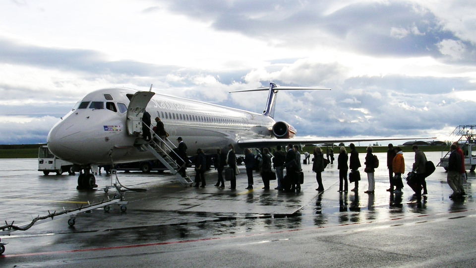 A smeller aircraft with a queue of persons waiting to board outdoors. Cloudy sky, wet ground.