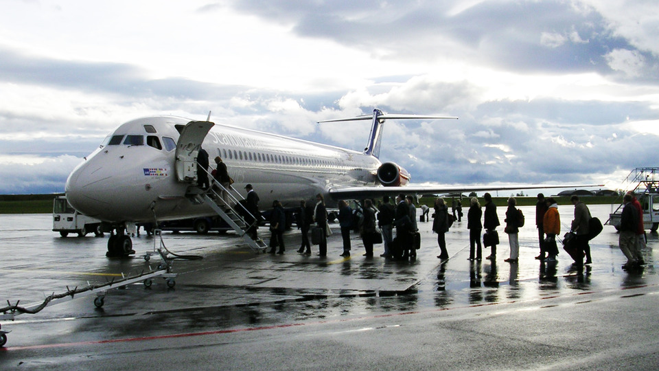 A smeller aircraft with a queue of persons waiting to board outdoors. Cloudy sky, wet ground.