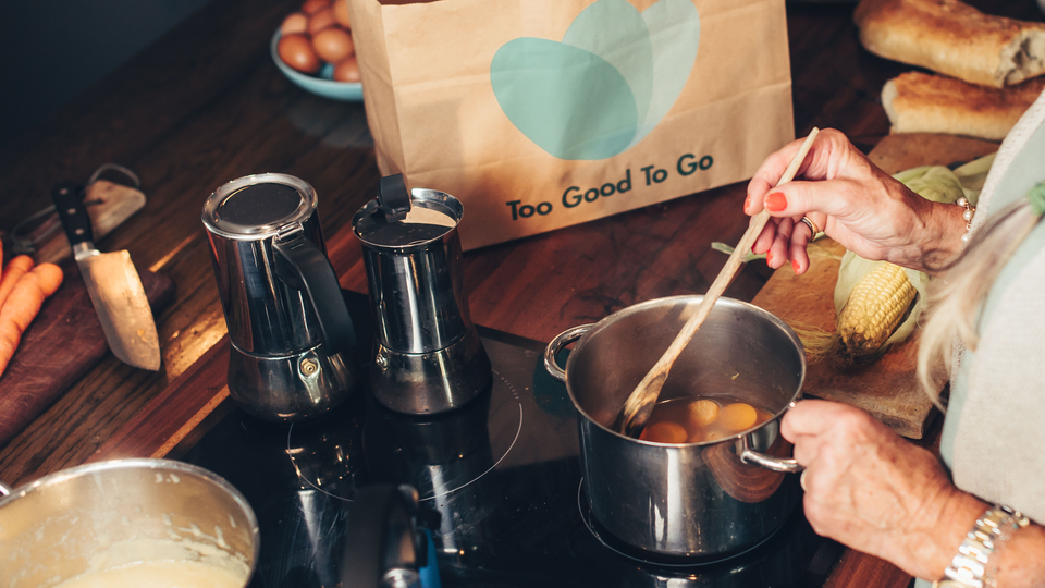 Close up of hands stirring in a pot on a stove, with a bag from ToGoodToGo in the background.