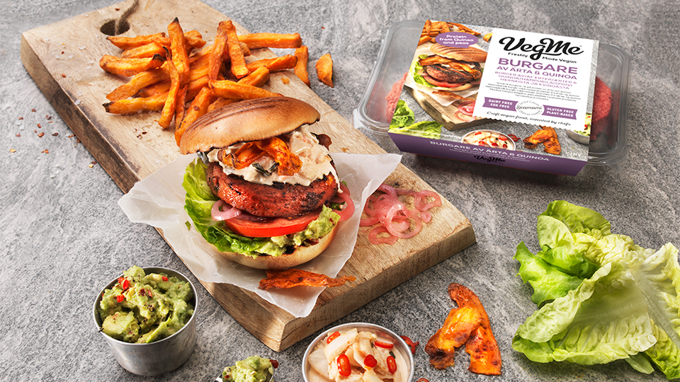 Press image of a plant-based burger lying on a cutting board with salad, french fries and other ingredients around it.