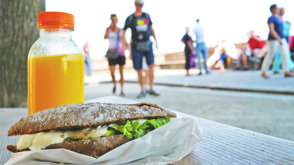 In the foreground: a sandwich and a bottle of juice, in the background: people moving in a city.