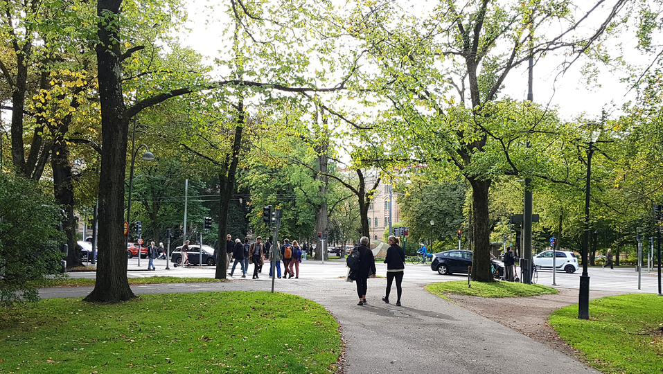 En asfalterad gångväg under stora träd som mynnar vid en vägkorsning. Personer som promenerar, några står vid ett övergångsställe.