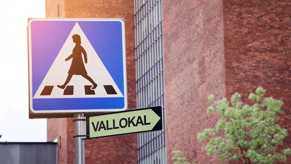 Direction sign in Swedish showcasing where the nearby polling station is located in the morning on election day in Sweden. Sign under pedestrian crossing sign in the city. Buildings in the background.