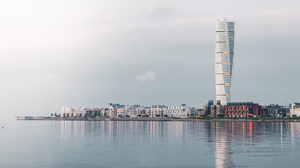A city silouette from Malmö with the tall building Turning Torso rising above the over houses. Calm sea in the front, slightly cloudy sky above. 