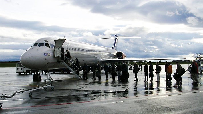 A smeller aircraft with a queue of persons waiting to board outdoors. Cloudy sky, wet ground.