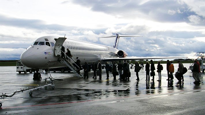 A smeller aircraft with a queue of persons waiting to board outdoors. Cloudy sky, wet ground.